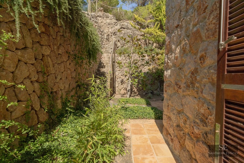 Terracotta tile path between rustic stone walls and lush greenery leading to stone stairs.
