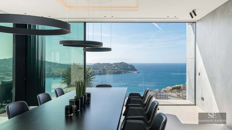 Modern black dining table and chairs facing floor-to-ceiling glass walls overlooking the blue ocean and coastline.