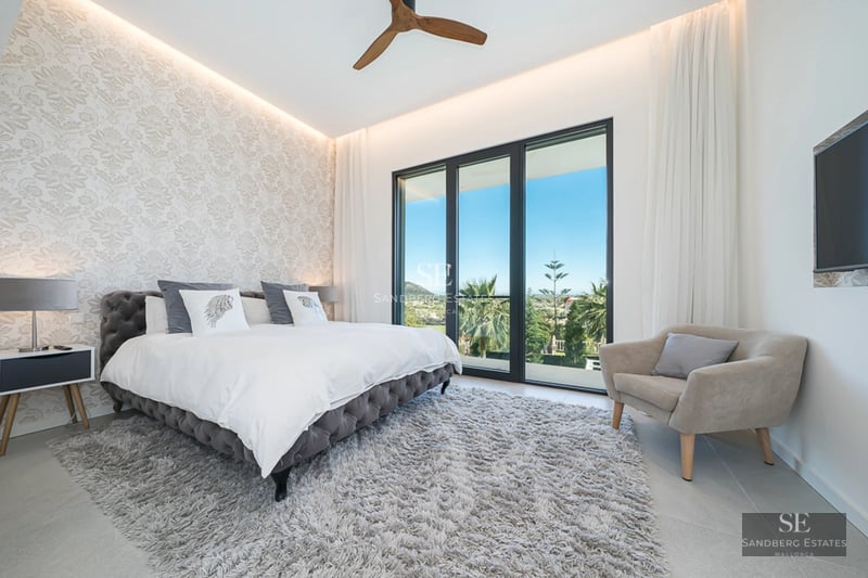 Panoramic view from a balcony with glass railing overlooking the sea. Modern and bright interior. Potted plants.