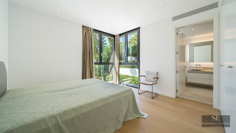 Bright master bedroom with wood floors, large corner windows overlooking greenery, and an adjacent modern bathroom.