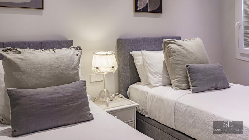 Two twin beds with grey upholstered headboards, white linens, and a lamp on a bedside table between them.