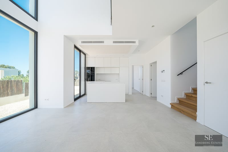Bright white minimalist kitchen with island, wooden stairs, and large floor-to-ceiling windows.