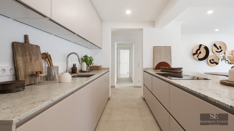 Modern kitchen with beige cabinets, stone countertops, and a view down a white hallway.