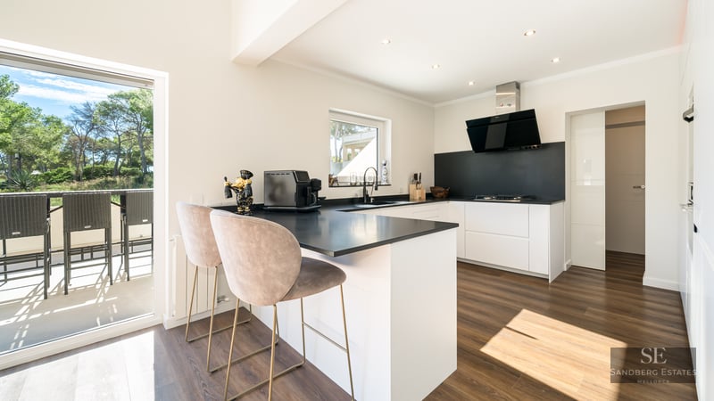 Cuisine moderne blanche avec plans de travail noirs, parquet et bar donnant sur une terrasse ensoleillée.