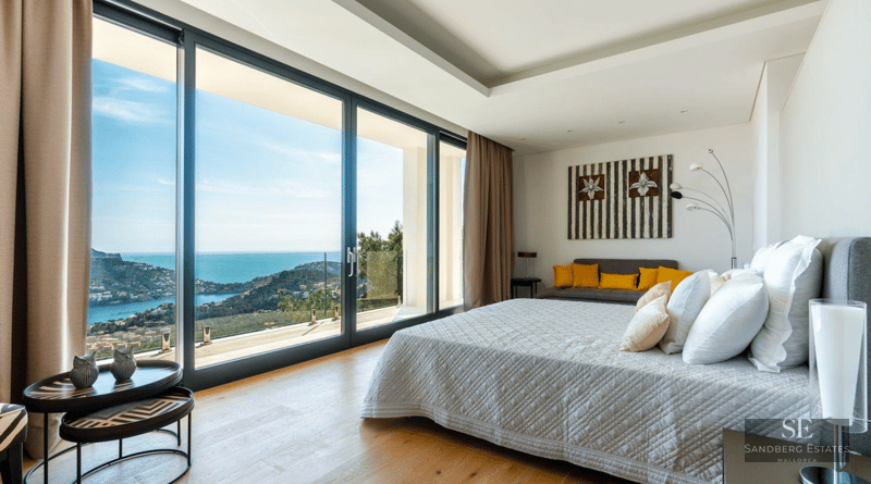 Modern master bedroom featuring floor-to-ceiling glass doors with a stunning view of the sea and coastline.