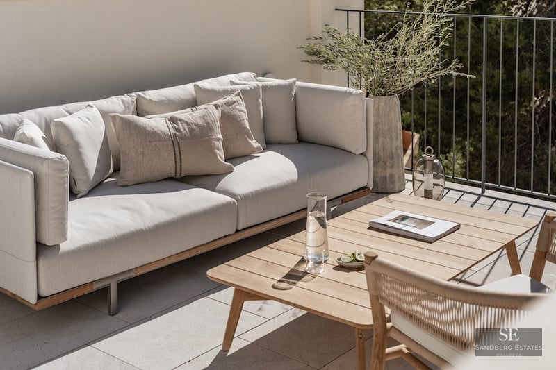 Modern outdoor terrace featuring a plush grey sofa, wooden coffee table, and greenery against a white wall and railing.