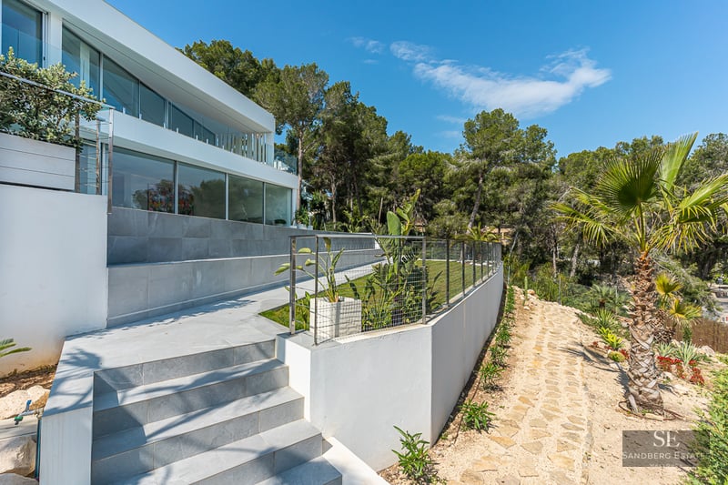 Modern white villa with a tiered garden featuring stone stairs, a lawn, and palm trees under a clear blue sky.