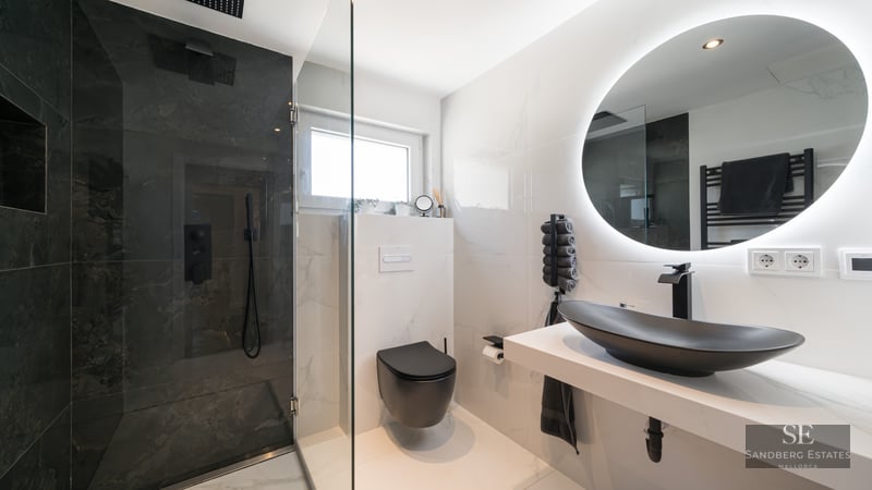 Salle de bain de luxe avec douche en marbre noir, murs en marbre blanc, vasque noir mat et miroir circulaire rétroéclairé.