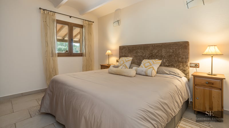 A cozy bedroom featuring a large bed with a brown velvet headboard, wooden nightstands, and warm ambient lamp lighting.