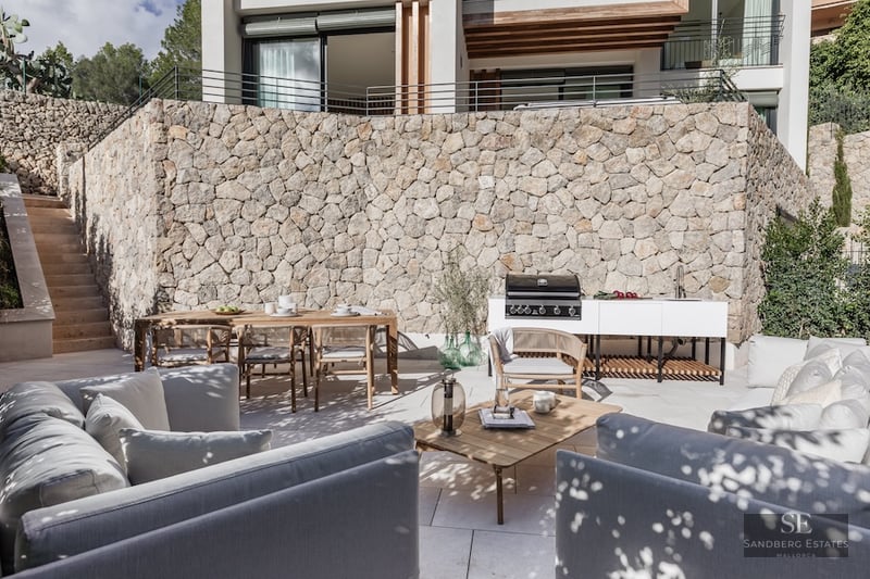 Modern terrace with a stone wall, outdoor kitchen, wooden dining table, and grey sofas under dappled natural light.