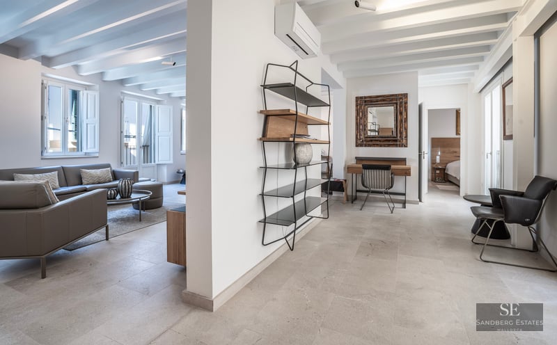 Grand salon moderne avec canapé en cuir gris, sols en pierre, poutres apparentes et étagère minimaliste.