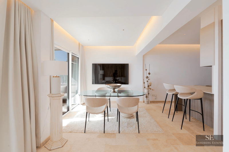 Minimalist dining room featuring a glass table, cream chairs, white rug, and a breakfast bar in the background.