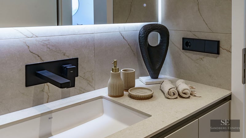 Close-up of a white sink with a matte black wall-mounted faucet, beige marble-effect tiling, and designer accessories.
