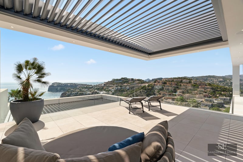 Modern terrace with stone tiling, sun loungers, a louvered pergola, and stunning views of the sea and coastal hills.