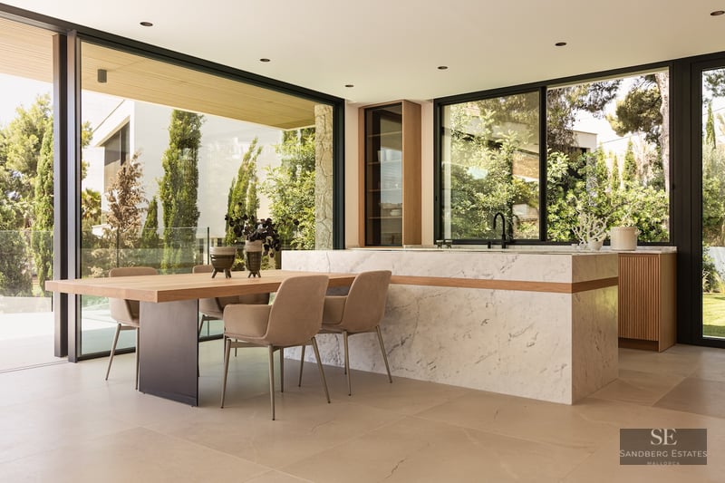 Modern kitchen with a massive marble island, wooden dining table, and floor-to-ceiling windows showing a lush garden.