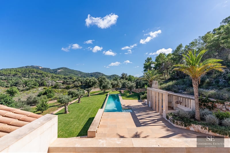 High-angle view of a long turquoise lap pool, stone terrace, and olive trees set against lush green hills and a blue sky.