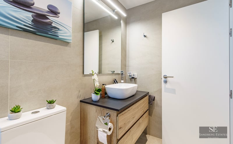 Modern bathroom featuring a white vessel sink on a wooden vanity, beige tiled walls, and a large mirror with LED light.