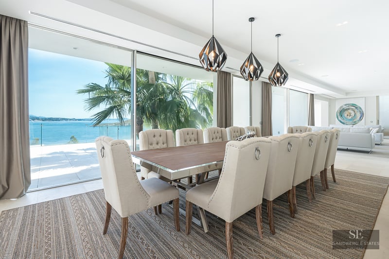 Elegant dining room featuring a long table, beige tufted chairs, and large glass doors opening to a sea view with palm trees.