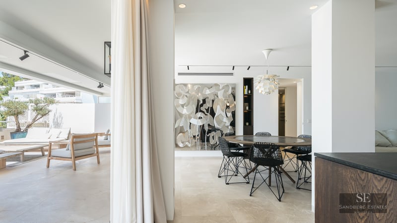 Modern indoor dining area with round wooden table opening onto a bright sun-drenched terrace with lounge seating.