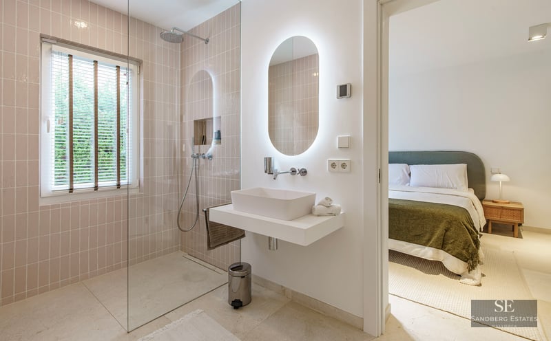 Modern bathroom with walk-in shower, backlit oval mirror, and view into a cozy bedroom.