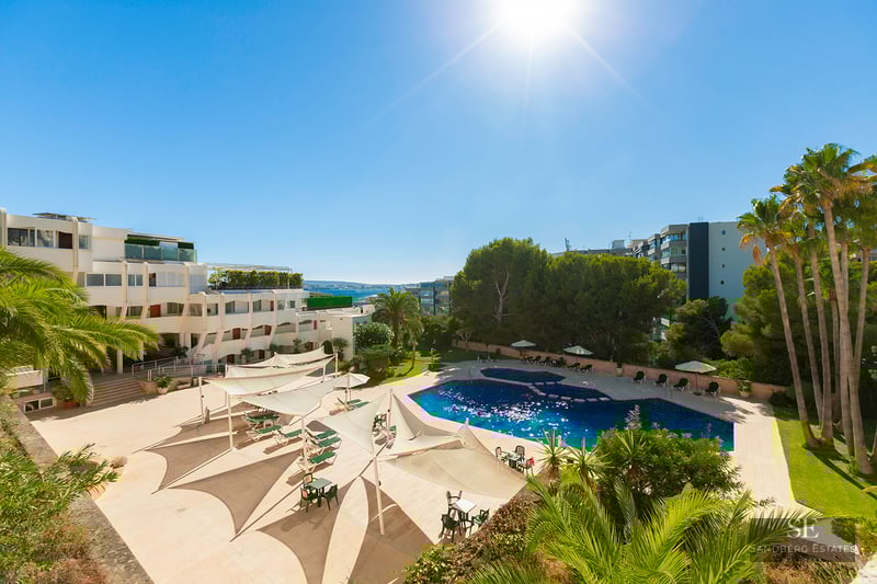 Large blue swimming pool surrounded by beige terrace, white sun sails, palm trees, and resort buildings under a bright sun.