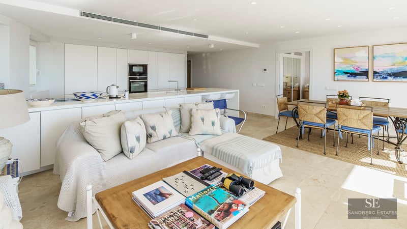 Bright open-plan living room with white sofa, wooden coffee table, dining set, and minimalist kitchen in the background.