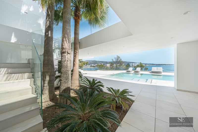 Modern villa terrace featuring a pool, palm trees, and glass railings overlooking the blue Mediterranean sea.