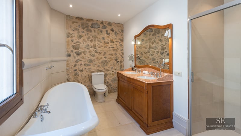 Elegant bathroom with a rustic stone wall, double wooden vanity, freestanding tub, and glass shower enclosure.