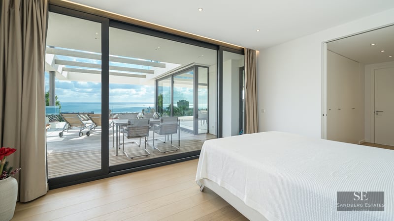 Modern bedroom with white bed and large glass doors opening to a terrace with sea views and outdoor furniture.