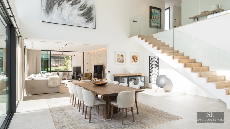 Salle à manger moderne avec table en bois, chaises blanches, escalier en verre et hauts plafonds lumineux.