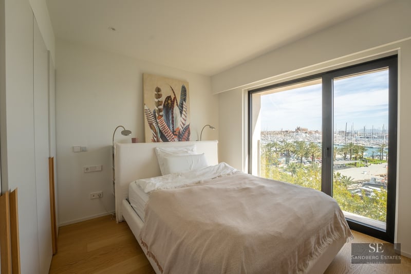 Modern bedroom with a white bed and a large window overlooking a sunny marina with palm trees.