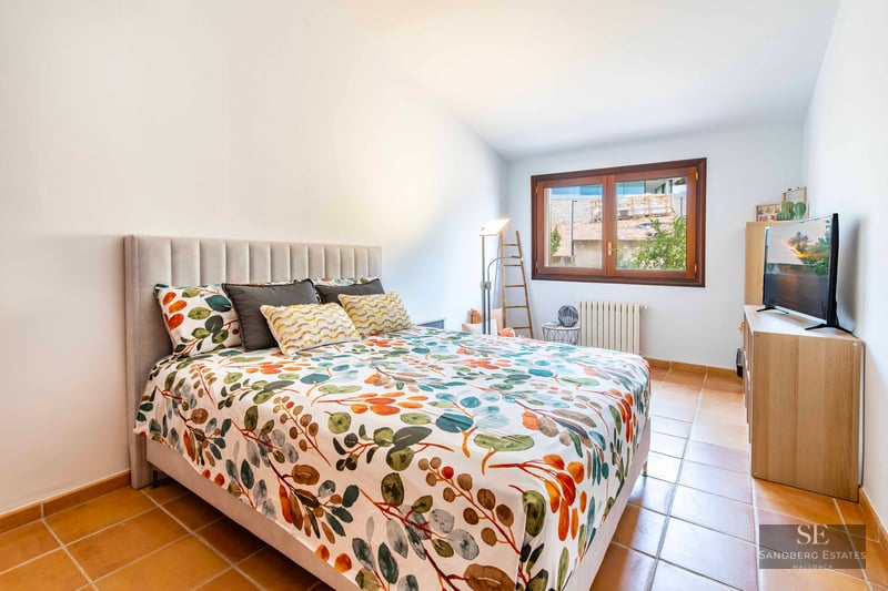 A bedroom featuring floral bedding, a grey upholstered headboard, terracotta tile floors, and a wooden window.