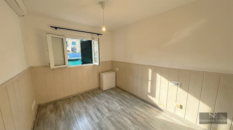 Empty room with light wood floors, partial wall paneling, an open window with green shutters, and a small white dresser.