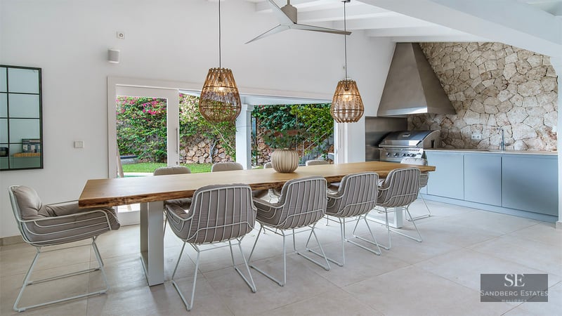 Large live-edge wooden dining table with designer chairs and woven pendant lights next to a stone-walled outdoor kitchen.