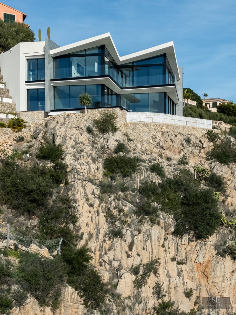 Modern white villa with large glass walls built on a steep rocky cliffside under a clear blue sky.