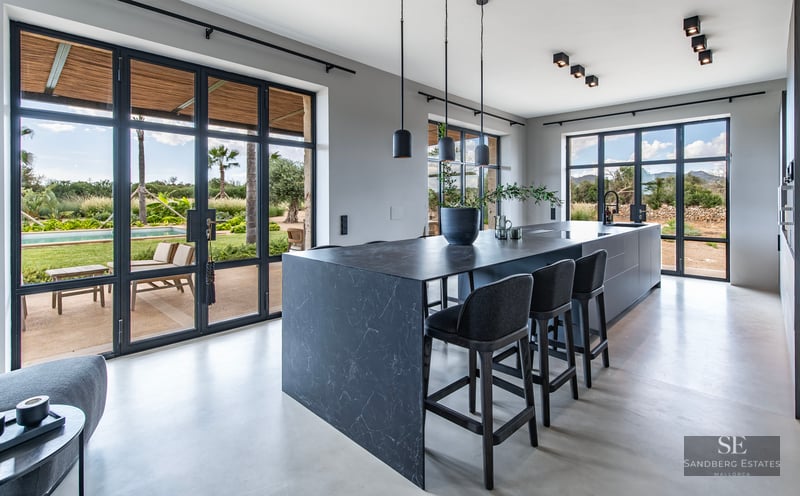 Modern kitchen featuring a large dark marble island, black stools, and glass doors opening to a garden and pool.