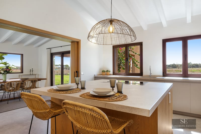 Modern kitchen island with wicker stools and white beamed ceiling overlooking rural landscape.