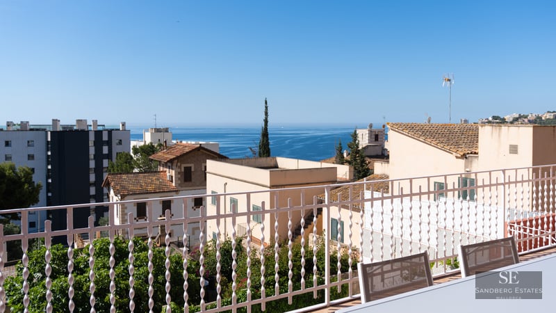 Panoramic view of the blue sea over coastal rooftops from a white balcony with outdoor seating.