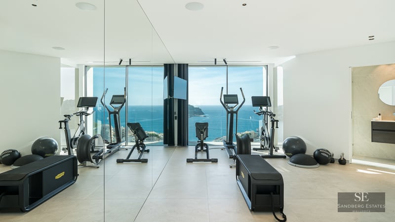 Modern home gym with professional equipment and floor-to-ceiling windows overlooking the blue Mediterranean sea.