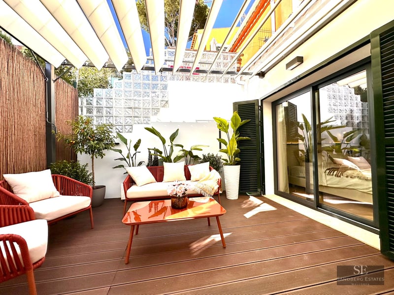 Stylish outdoor terrace with red and white furniture, orange table, lush plants, and a white retractable awning in the sun.