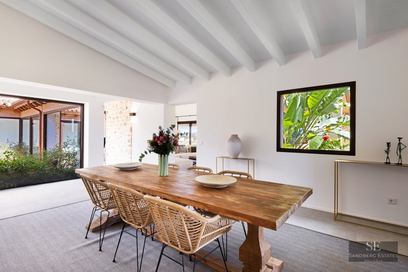Bright dining room with a large wooden table, wicker chairs, white beamed ceiling, and lush garden views.