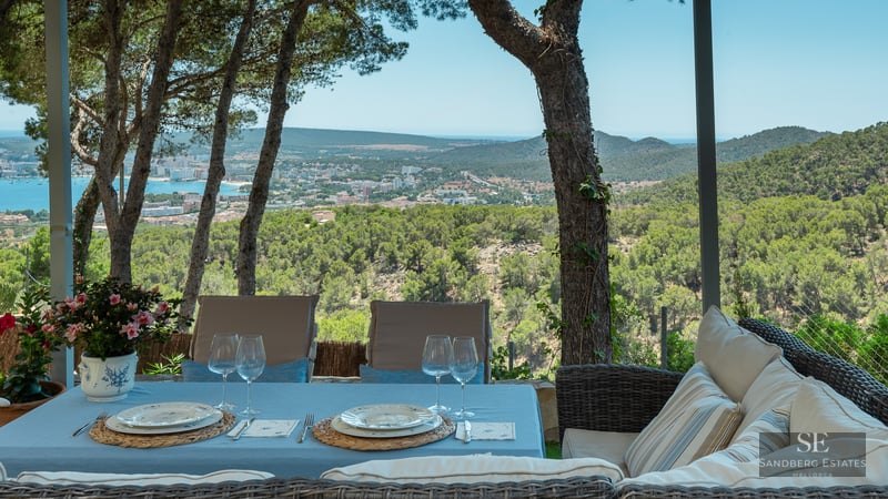 Outdoor dining table and wicker sofa on a shaded terrace overlooking a coastal town and sea.