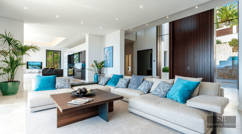 Spacious white living room with a large gray sectional sofa, teal pillows, dark wood accents, and an open kitchen view.