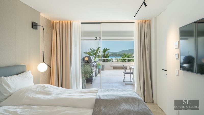 Modern bedroom with a white bed and large glass doors opening to a terrace with a BBQ and sea views.