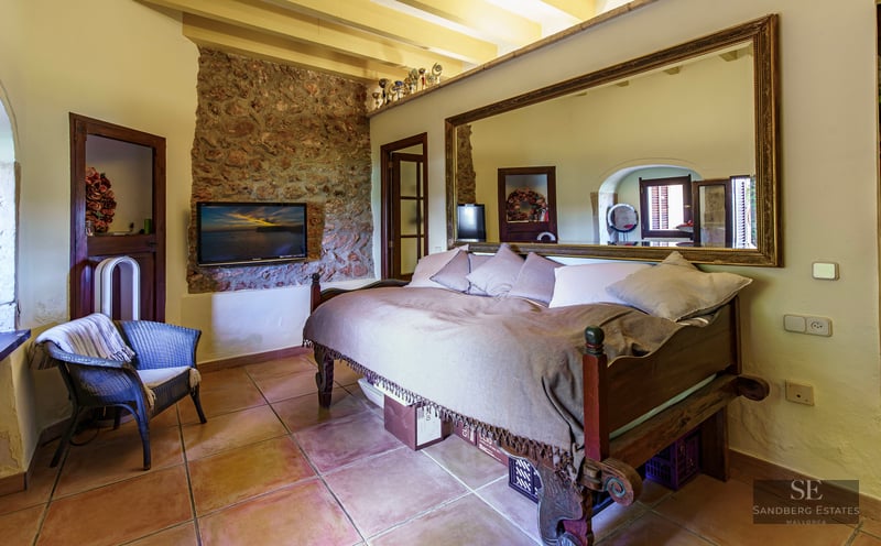 A cozy bedroom featuring terracotta tile flooring, a natural stone wall with a TV, and a large ornate mirror above the bed.
