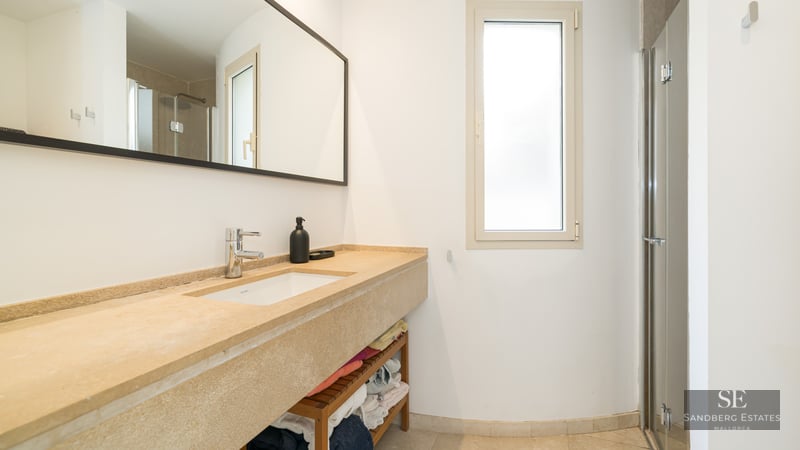 Modern bathroom featuring a long beige stone vanity, large black-framed mirror, and glass shower door in a bright room.