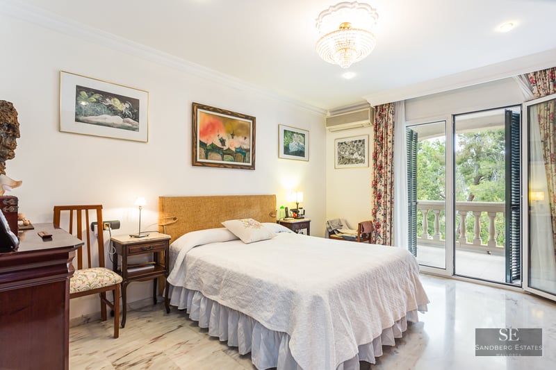 Elegant bedroom with a double bed, marble flooring, crystal chandelier, and glass doors opening to a balcony.