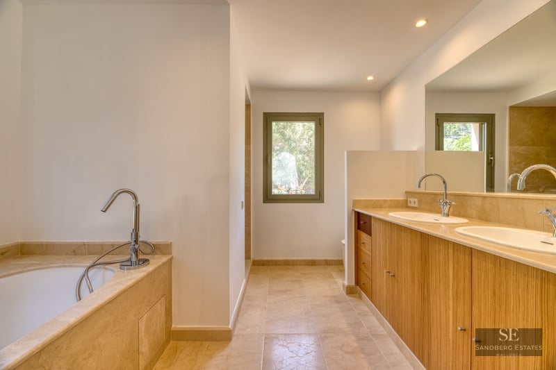 Luxury bathroom featuring wooden double vanity, beige marble countertops, bathtub, and natural light.