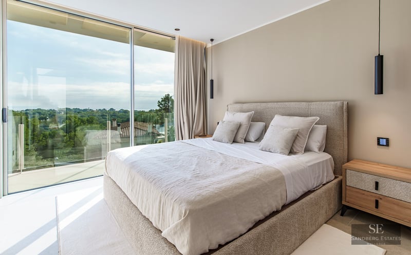Modern master bedroom featuring floor-to-ceiling glass walls overlooking a lush green landscape with a large neutral bed.