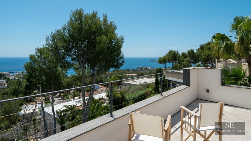 Sunny terrace with metal railing, two wooden chairs, and panoramic views of the blue sea and green pine trees.